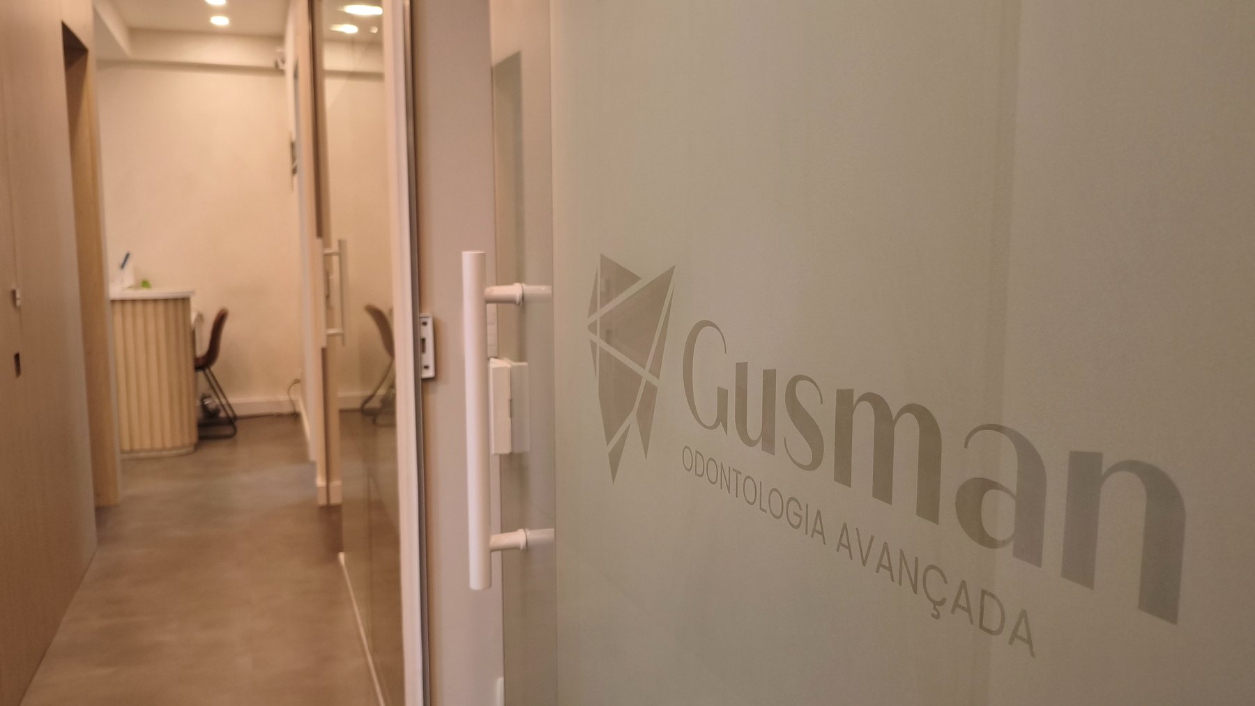 Entrada da Clínica Gusman, com o logotipo aplicado na porta de vidro jateado e o corredor de consultórios ao fundo
